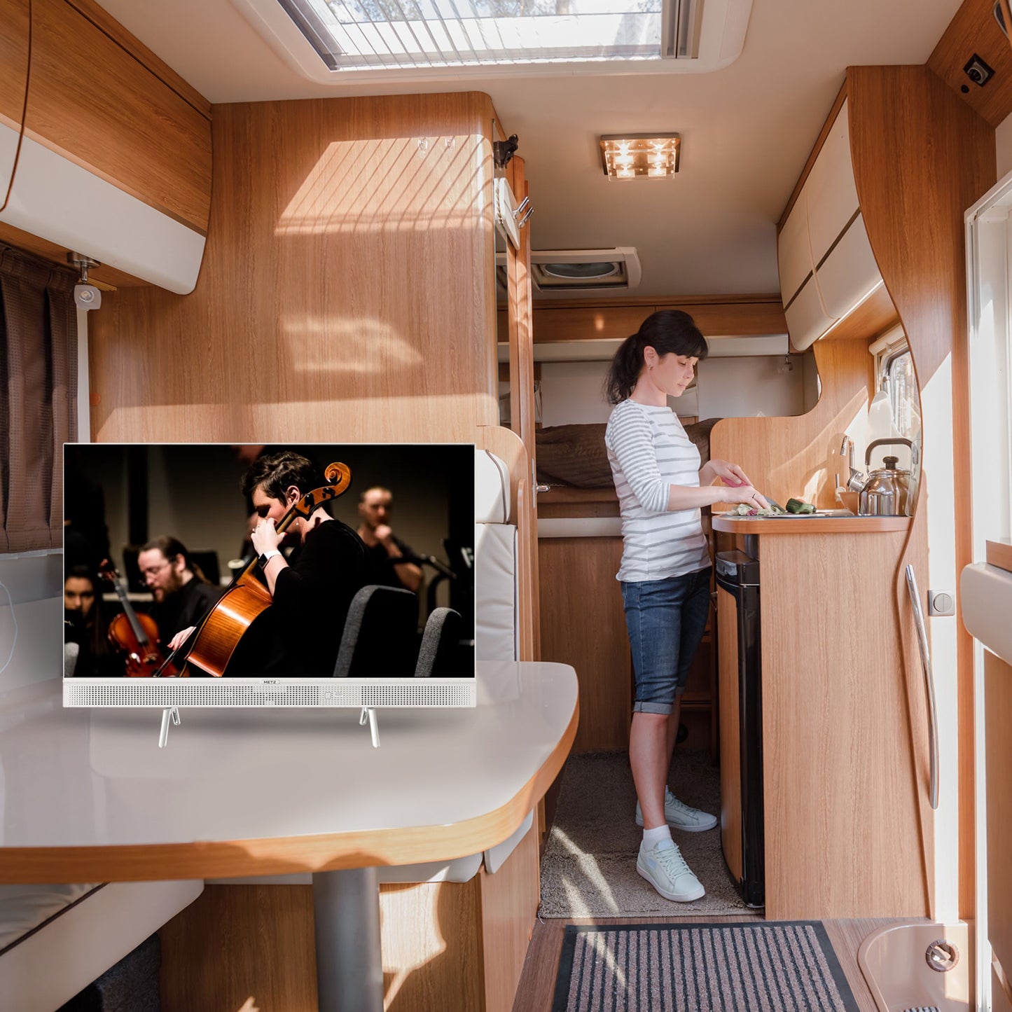 Metz Bærbar 12 V Google TV 32" på bord i campingkøkken, viser orkester med cellospiller, træpaneler og ovnlys.
