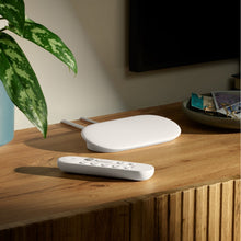 Google TV-streamer 4K Bundle med stemmestyret fjernbetjening på træskab, grøn plante og nøglebakke ved væghængt TV.
