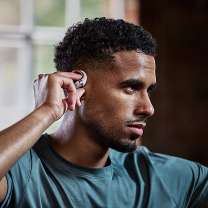 Shokz OpenFit 2+ TWS øretelefoner Sort tages på af mand i turkis skjorte, brandlogo synligt, sløret vindue baggrund.