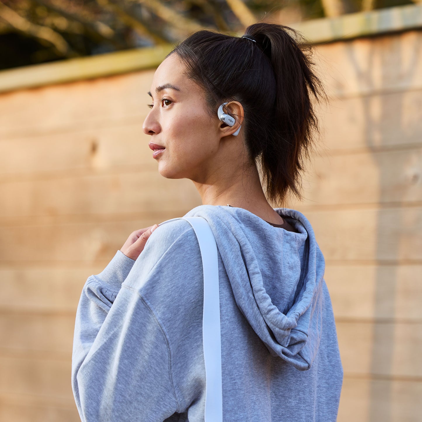 Shokz OpenFit 2+ TWS øretelefoner Sort bæres af kvinde med grå hættetrøje udendørs, taske over skulderen.