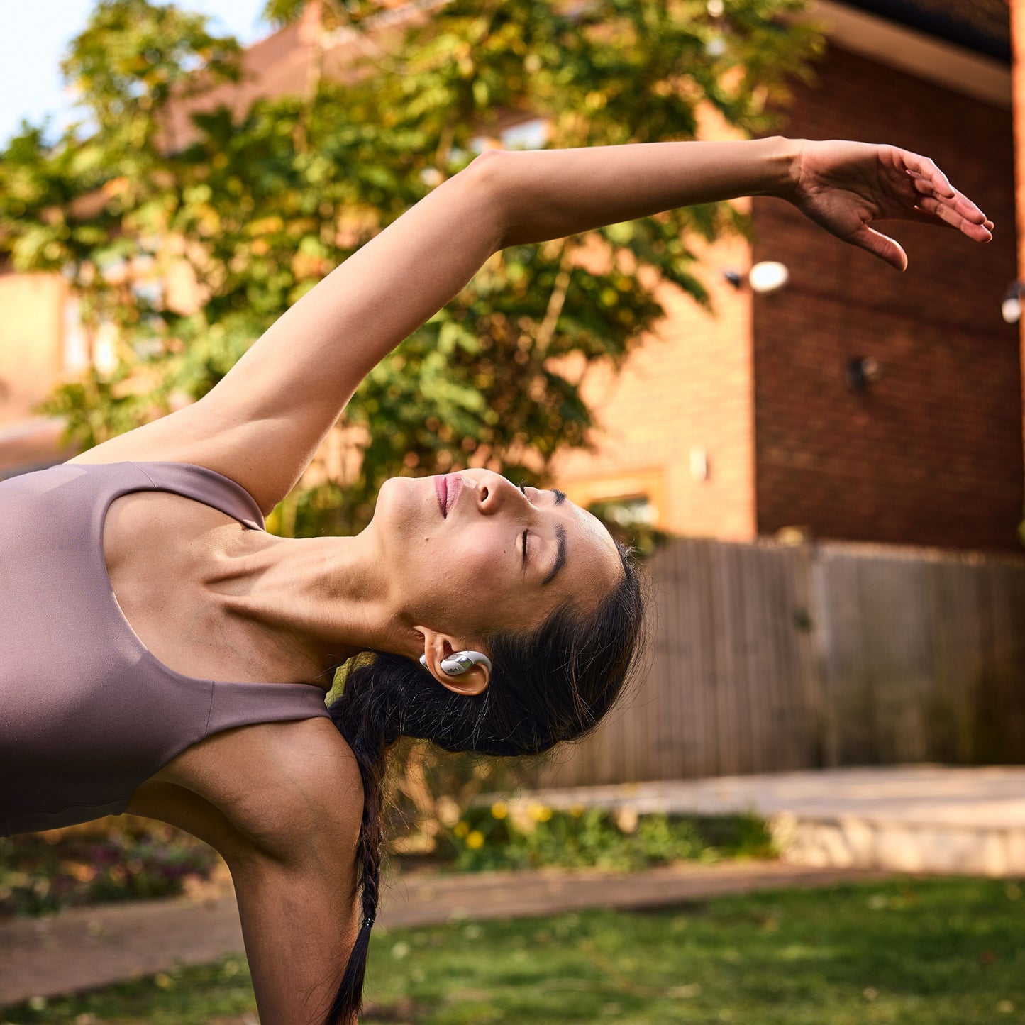 Shokz OpenFit 2+ TWS øretelefoner Sort bæres af kvinde iført ærmeløs top udendørs med træer og hus i baggrunden.