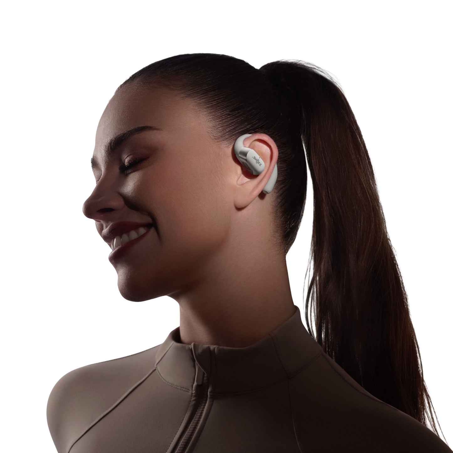 Shokz OpenFit 2+ TWS øretelefoner Grå på kvinde med langt mørkt hår i hestehale og højhalset beige top.