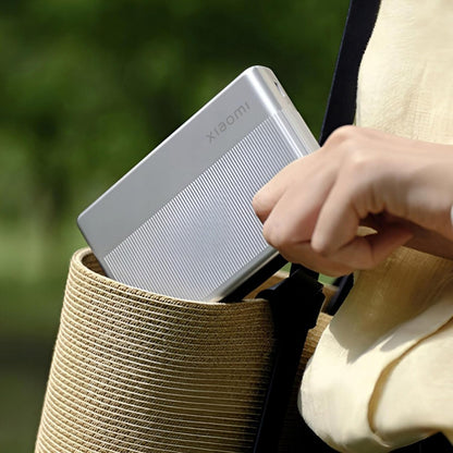 Xiaomi powerbank sølv ved siden af Xiaomi Bærbar fotoprinter 1S i stråtaske, sløret grøn baggrund.