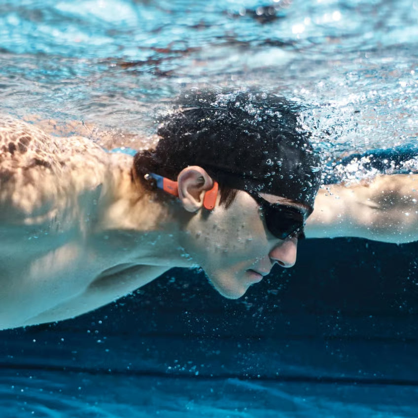 SHOKZ OpenSwim Pro trådløse hovedtelefoner – røde – på en svømmer med svømmebriller, badehætte og orange ørepropper under vandet.