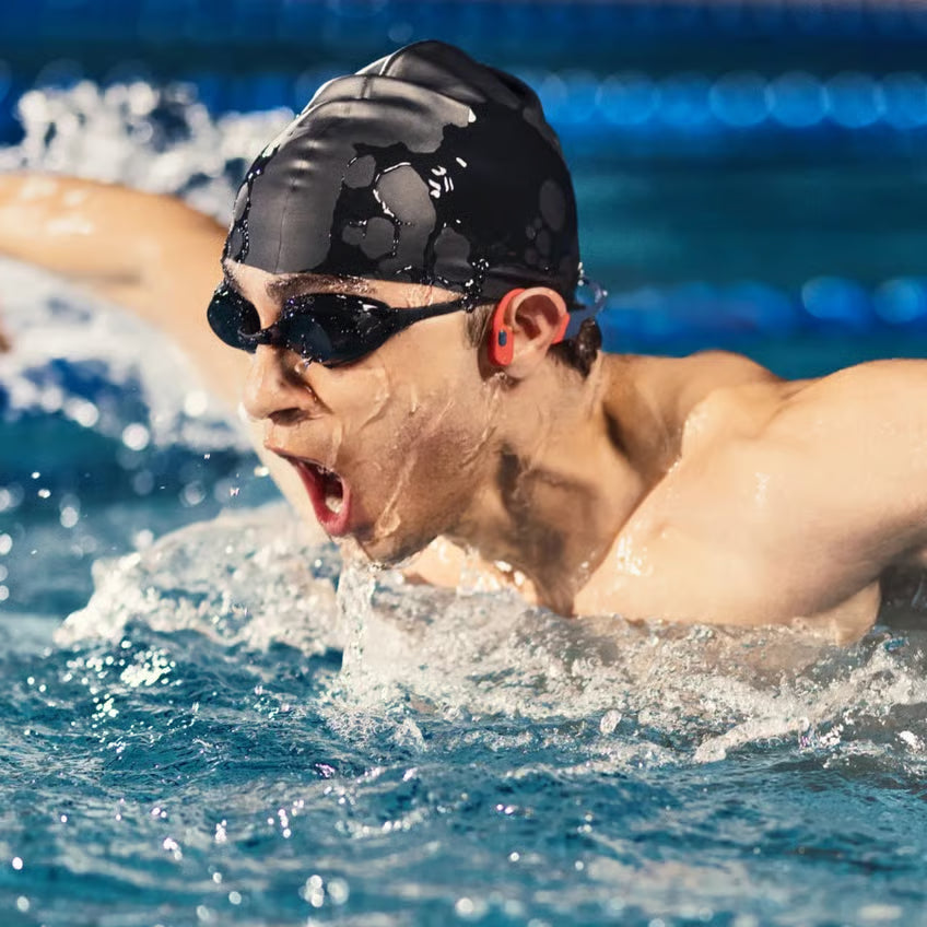 SHOKZ OpenSwim Pro trådløse hovedtelefoner (røde) bæres af en svømmer med svømmebriller og badehætte under butterfly i svømmehallen.