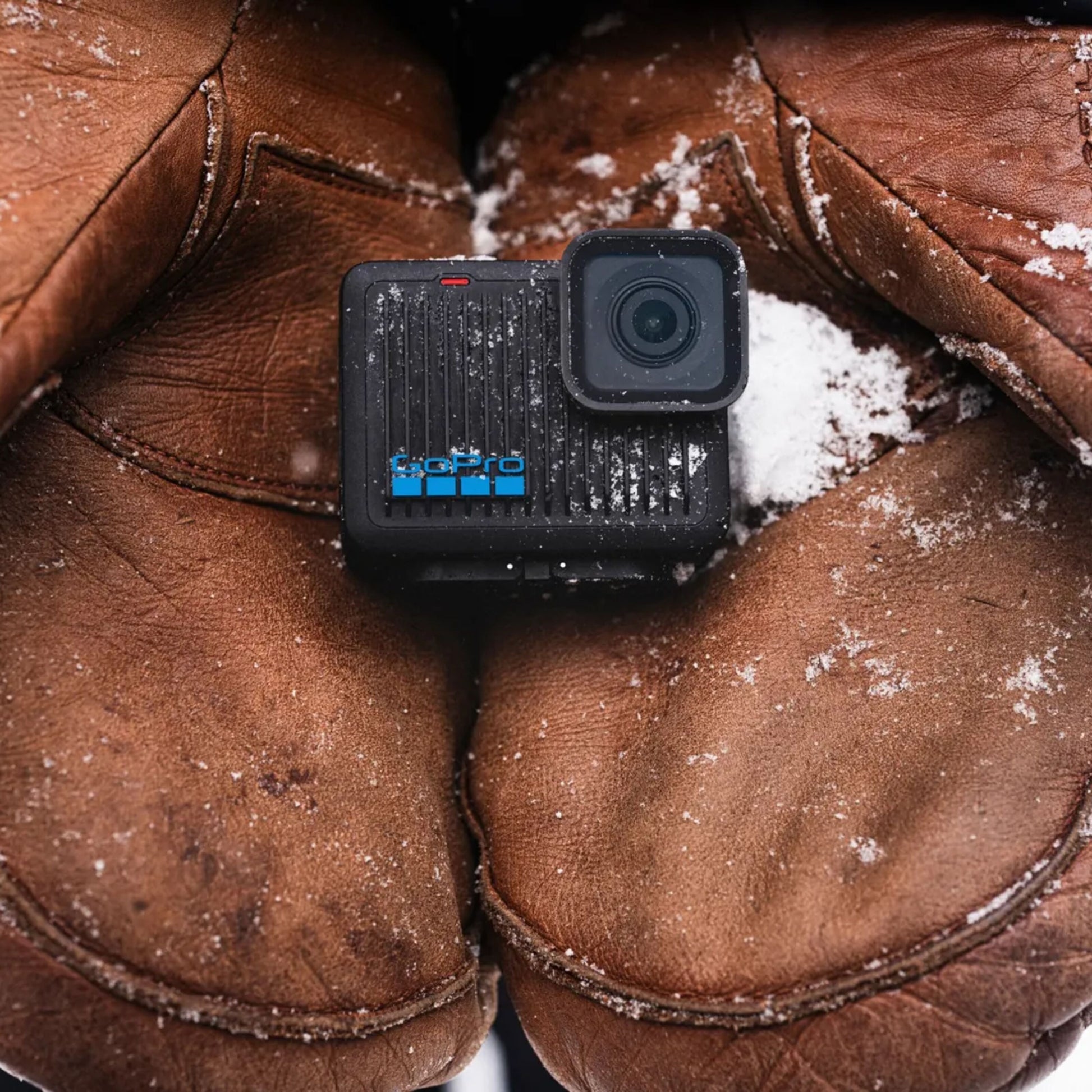 GoPro HERO actionkamera 4K mellem to brune læderhåndsker med sne, sort kompakt design, tydeligt brand.