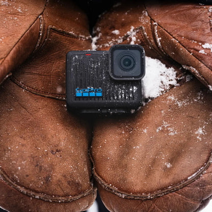 GoPro HERO actionkamera 4K mellem to brune læderhåndsker med sne, sort kompakt design, tydeligt brand.