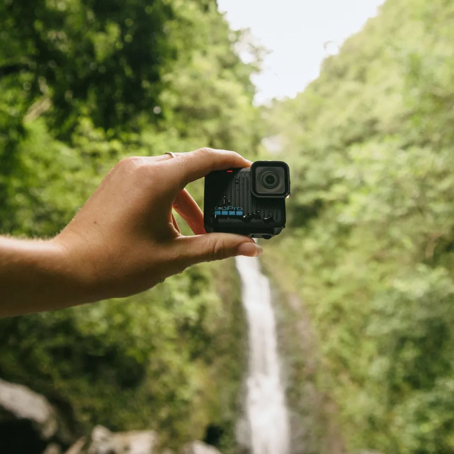 Gopro HERO actionkamera 4K holdes i hånden foran grøn skov og vandfald, sort kompakt design, linse synlig.
