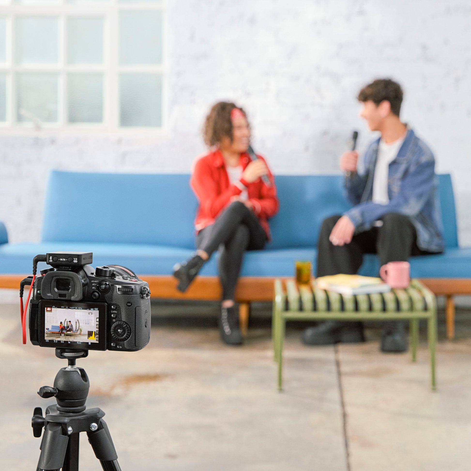 Røde Interview trådløs mikrofon med GainAssist i brug, blå sofa, stativ-kamera, kaffebord med krus og papirer.