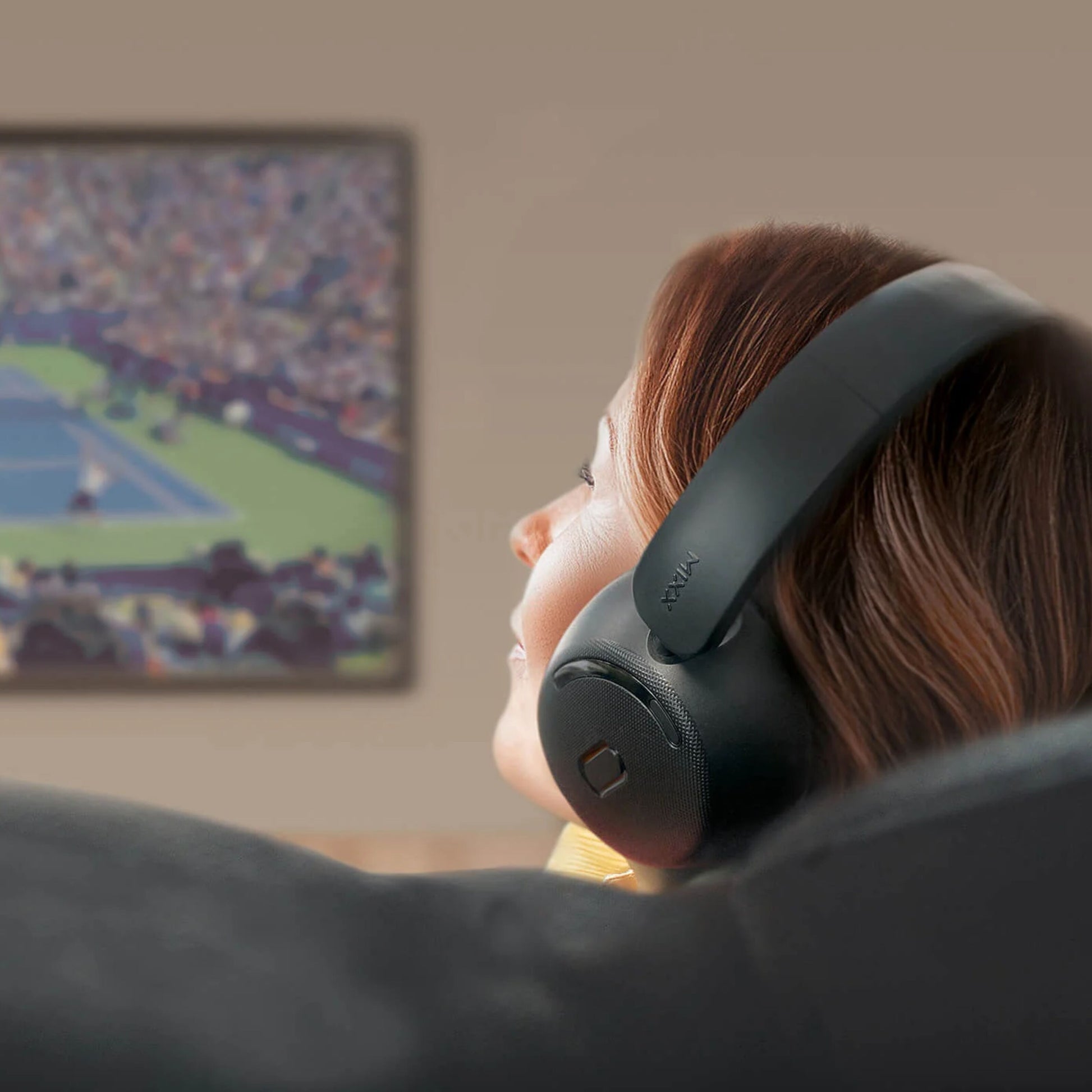 MIXX StreamQ Surround TV trådløse hovedtelefoner bæres af person i sofa foran vægmonteret tv-skærm med tenniskamp.
