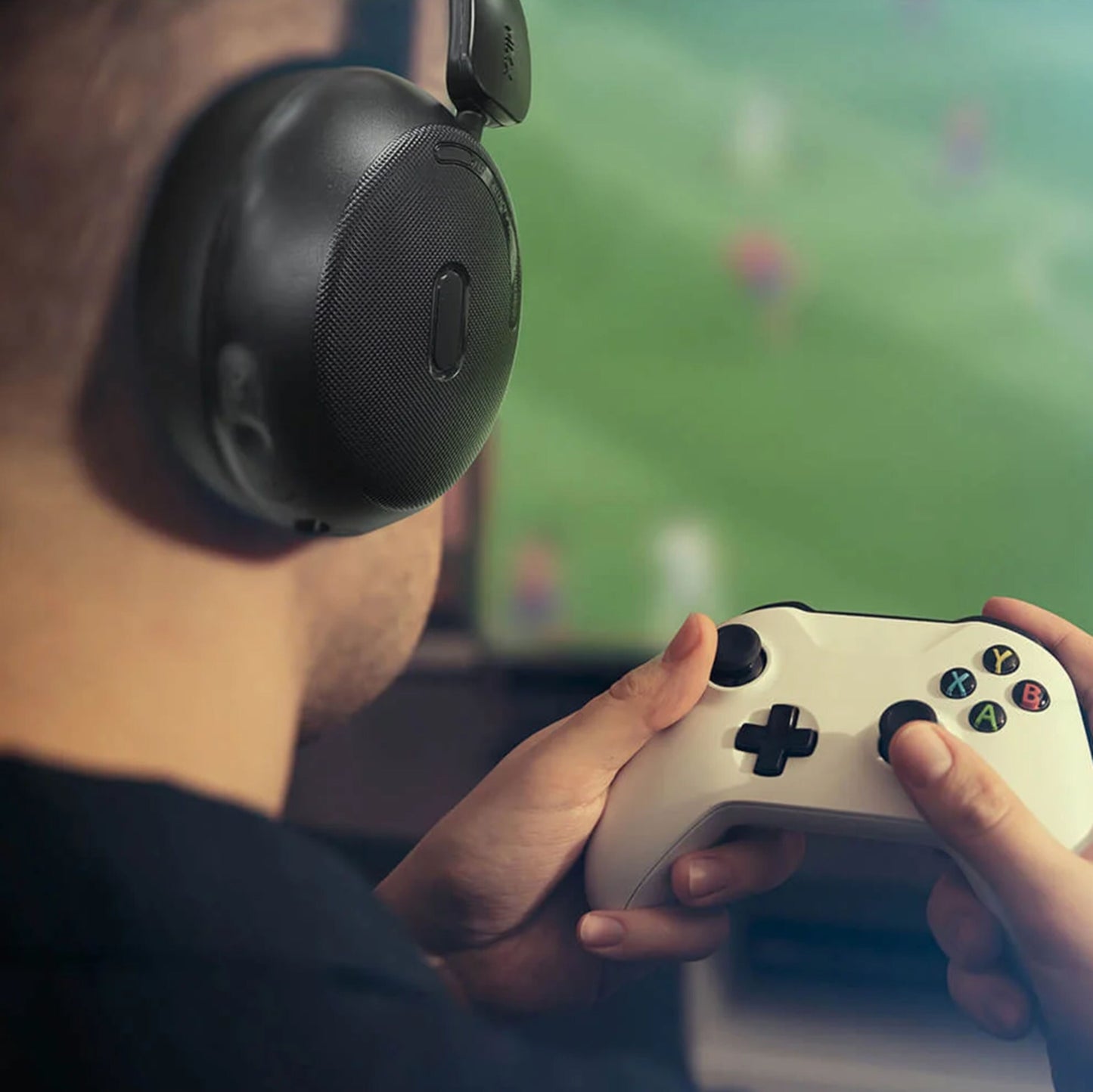 MIXX StreamQ Surround TV trådløse hovedtelefoner bæres af person med game controller, sløret sport på skærm i baggrund.