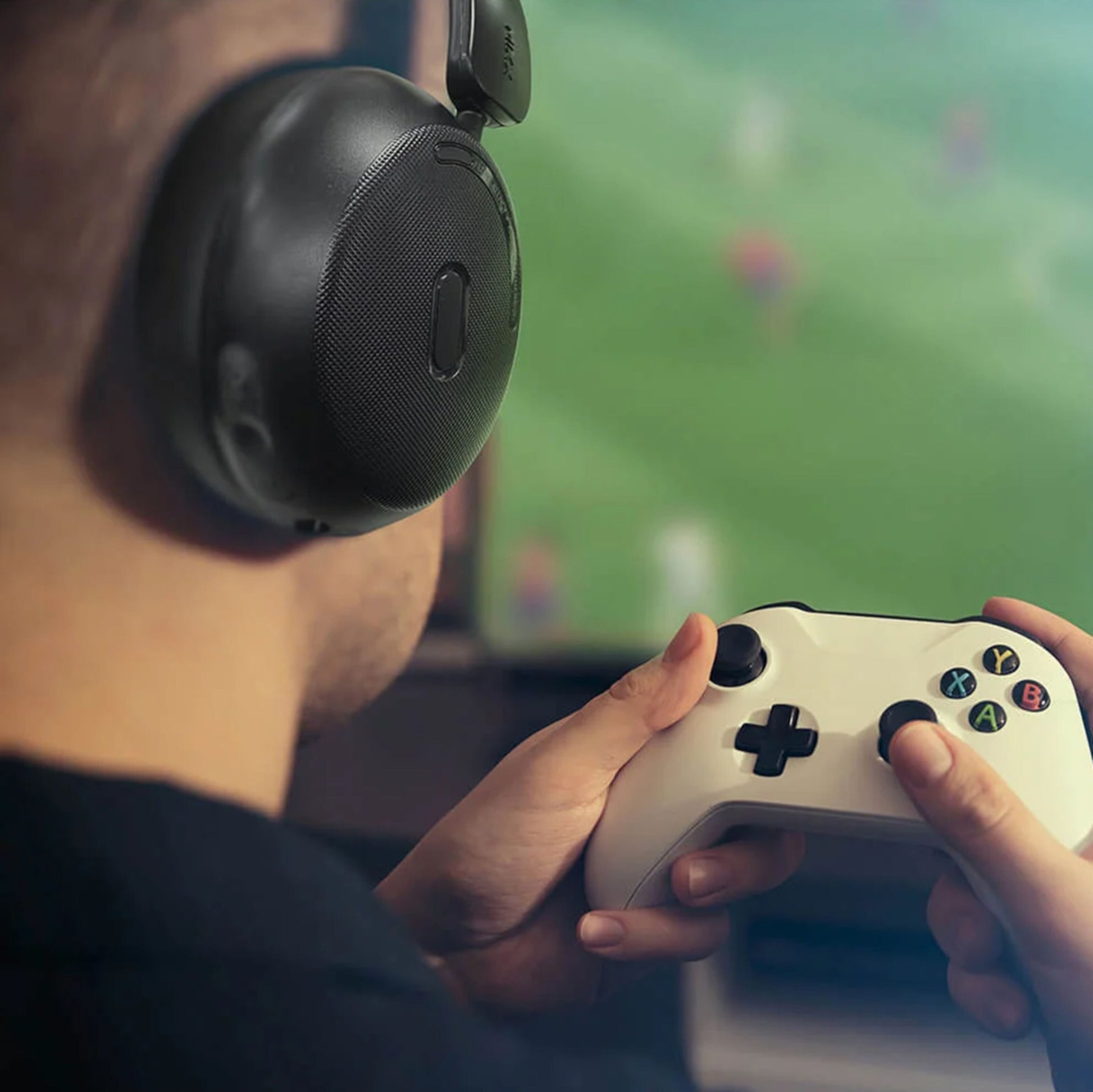 MIXX StreamQ Surround TV trådløse hovedtelefoner bæres af person med game controller, sløret sport på skærm i baggrund.
