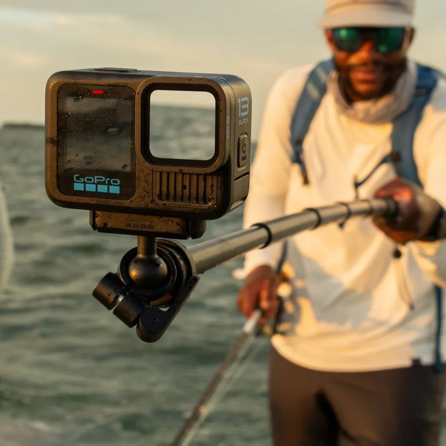 Gopro Magnetisk lås kugleled på forlængerstang, udendørs brug ved vand med person i fiskeudstyr i baggrunden.