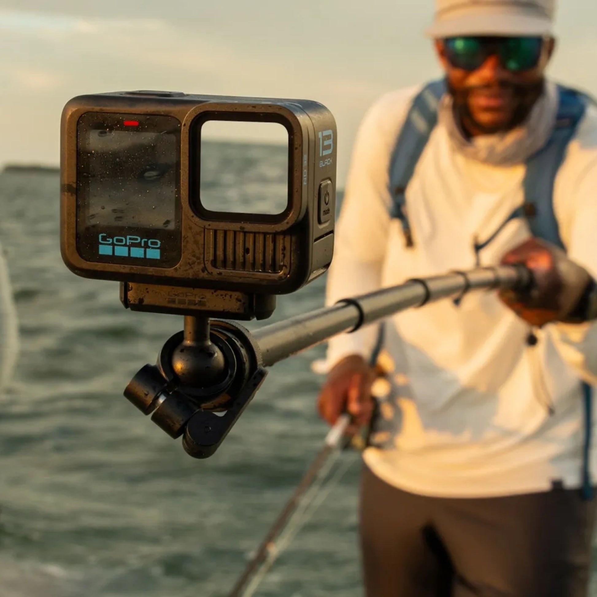 Gopro Magnetisk lås kugleled på forlængerstang, udendørs brug ved vand med person i fiskeudstyr i baggrunden.
