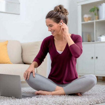 Hama Spirit Calm TWS øretelefoner sættes i øret af kvinde, laptop åben ved siden af, sofa og reol ses bagved.