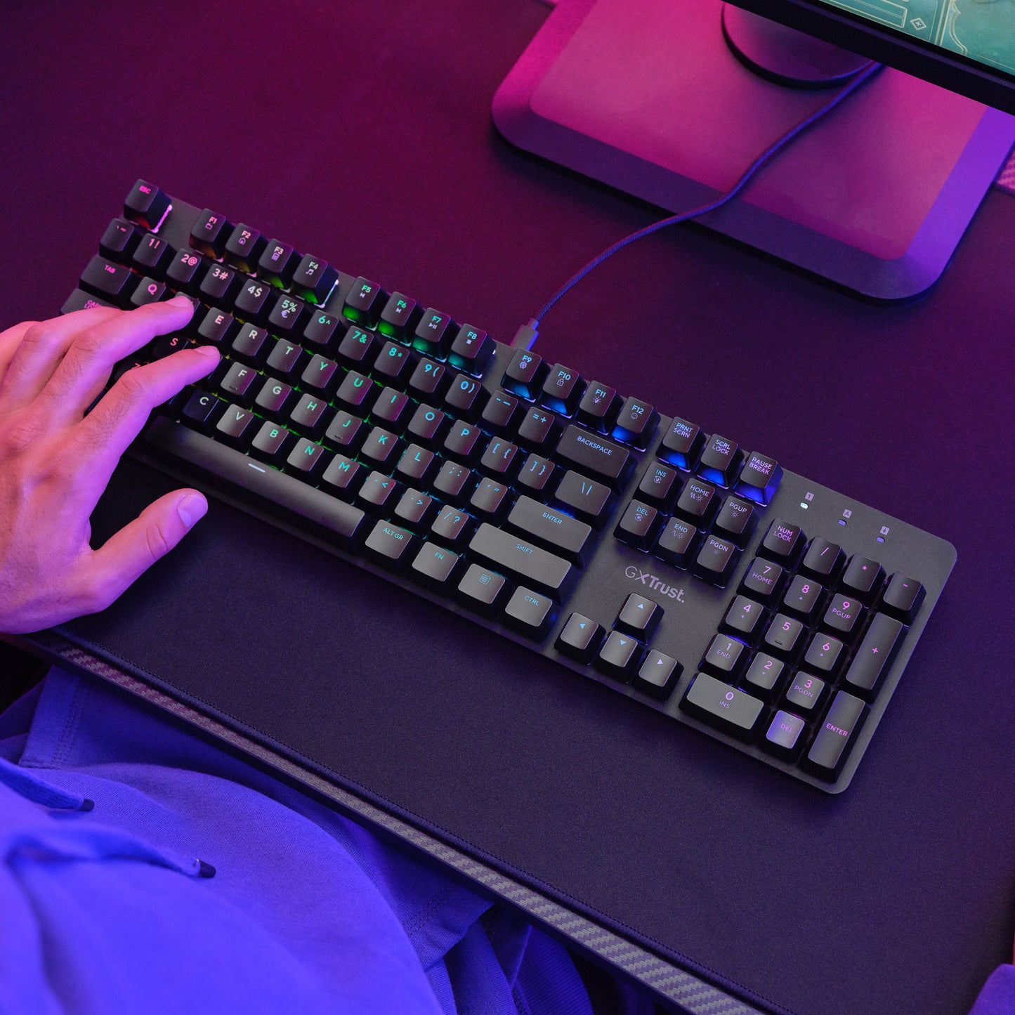 Trust GXT 871 Zora mekanisk gamingtastatur med RGB-belysning og Huano-switches på mørkt bord under lilla/pink lys.