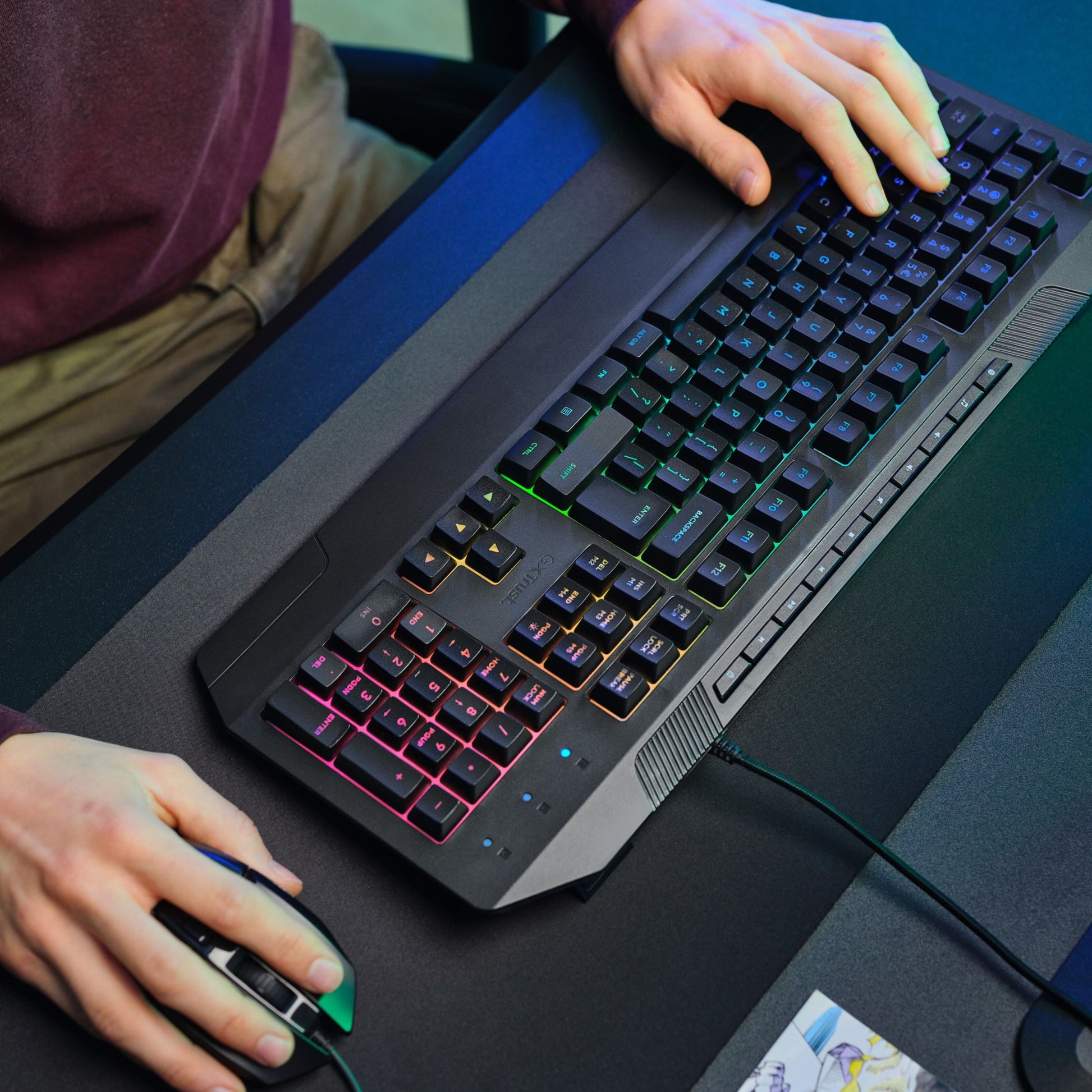 Trust GXT 836 Evocx RGB Gamingtastatur og computermus på bord, kun hænder og arme synlige.