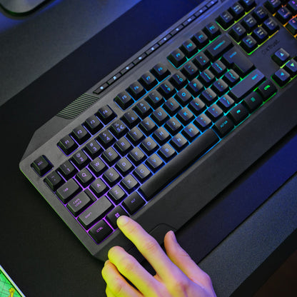 Trust GXT 836 Evocx RGB gamingtastatur med hånd på Enter-tasten, synlig RGB-belysning og anti-ghosting på mørkt skrivebord.