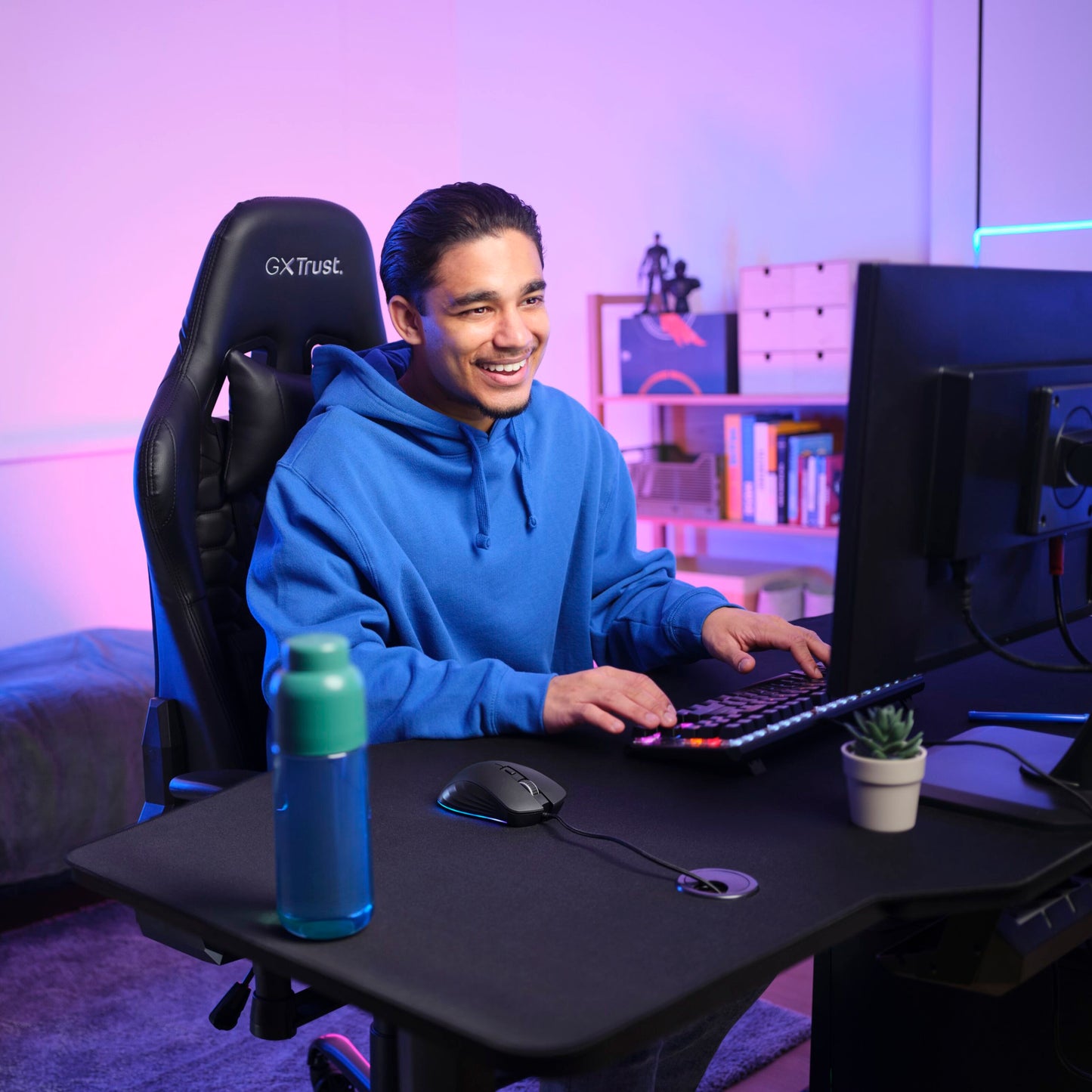 Trust GXT 924 Ybar+ gamingmus med PMW3389 sensor ved computer, synligt tastatur og blå/lilla belysning.