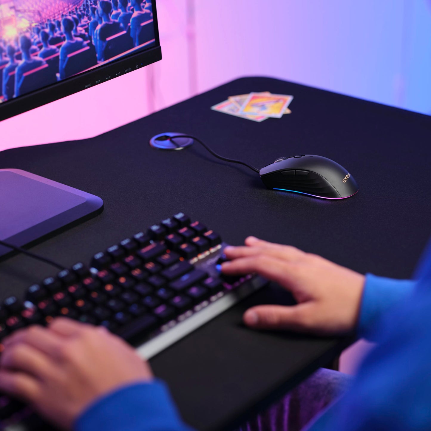 Trust GXT 924 Ybar+ gamingmus med PMW3389 sensor på skrivebord ved tastatur, kort og monitor med teaterscene, blå-pink lys.