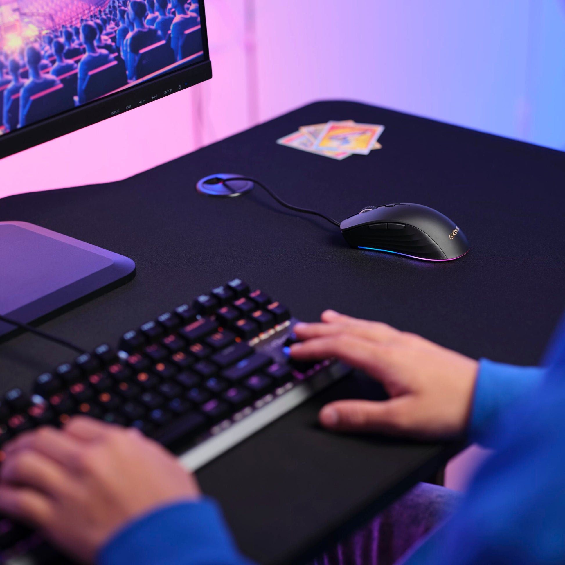 Trust GXT 924 Ybar+ gamingmus med PMW3389 sensor på skrivebord ved tastatur, kort og monitor med teaterscene, blå-pink lys.