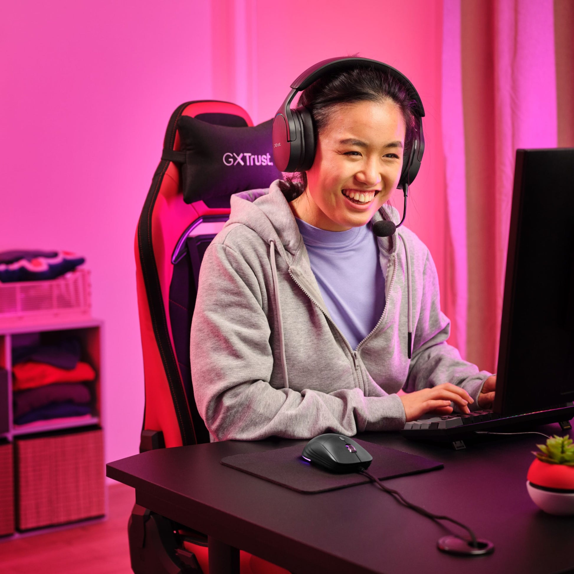 Trust GXT 925 Redex II gamingmus sort ses brugt ved sort skrivebord, pink RGB-lys, gamingstol synlig, kvinde iført grå hoodie.