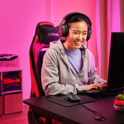 Trust GXT 925 Redex II gamingmus sort ses brugt ved sort skrivebord, pink RGB-lys, gamingstol synlig, kvinde iført grå hoodie.
