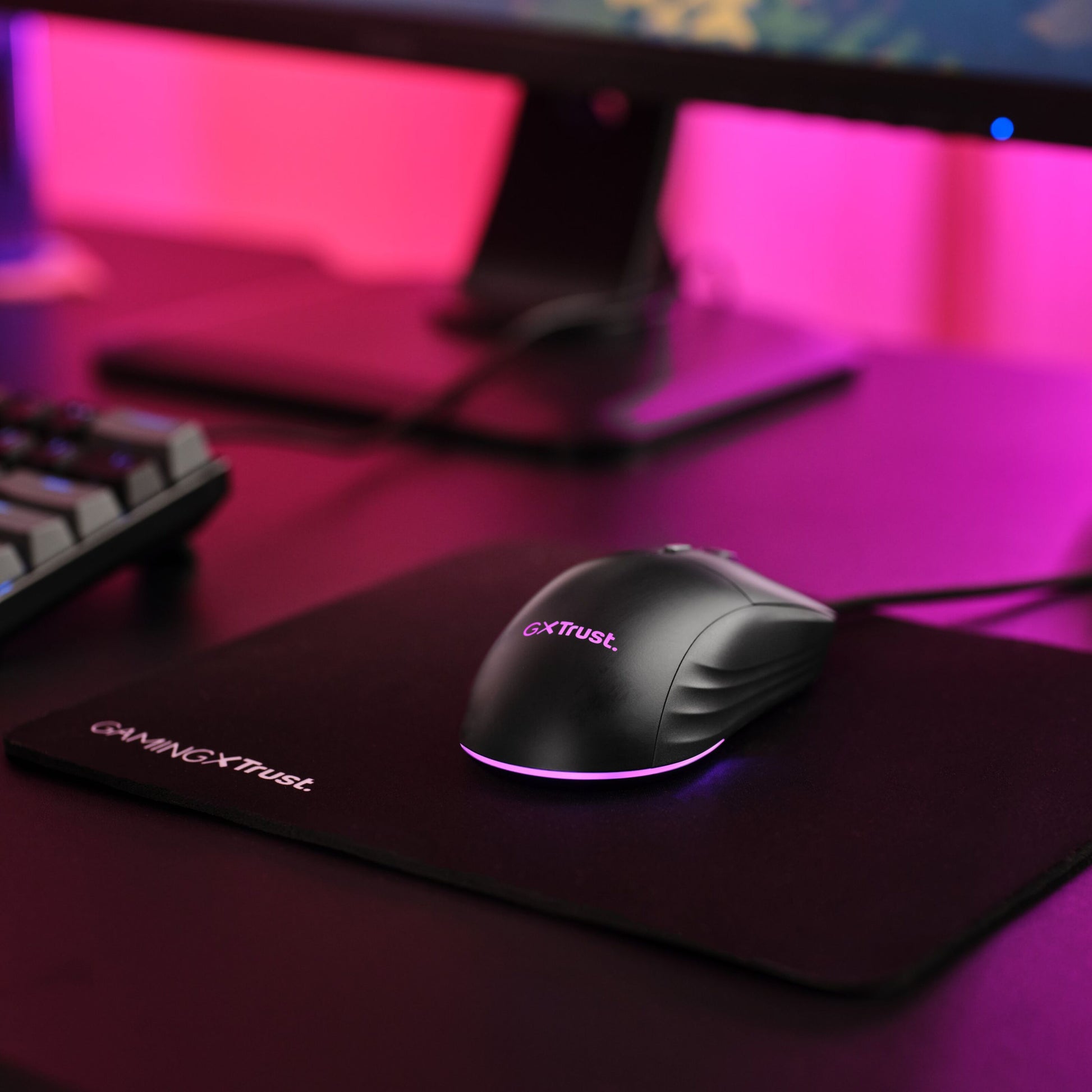 Trust GXT 925 Redex II gamingmus RGB-belysning på sort musemåtte ved siden af mekanisk tastatur og skærm med pink baglys.
