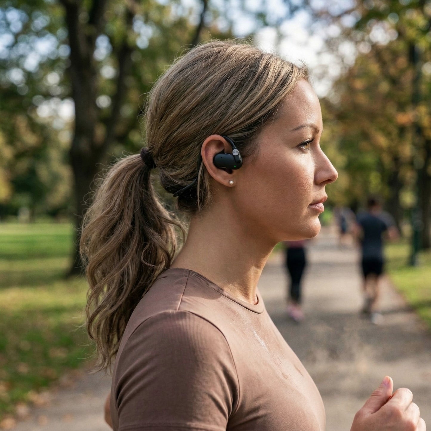 Nomadelic Solo 330 Sport open-ear-hovedtelefoner på kvinde med lysbrunt hår i hestehale og brun trøje i parkomgivelser.