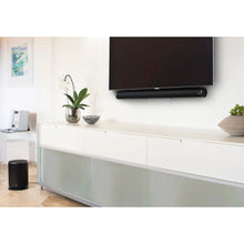 Hama Fleksibelt vægbeslag til soundbar monteret med sort soundbar under stor TV på hvid væg, konsol med hvide skuffer under.
