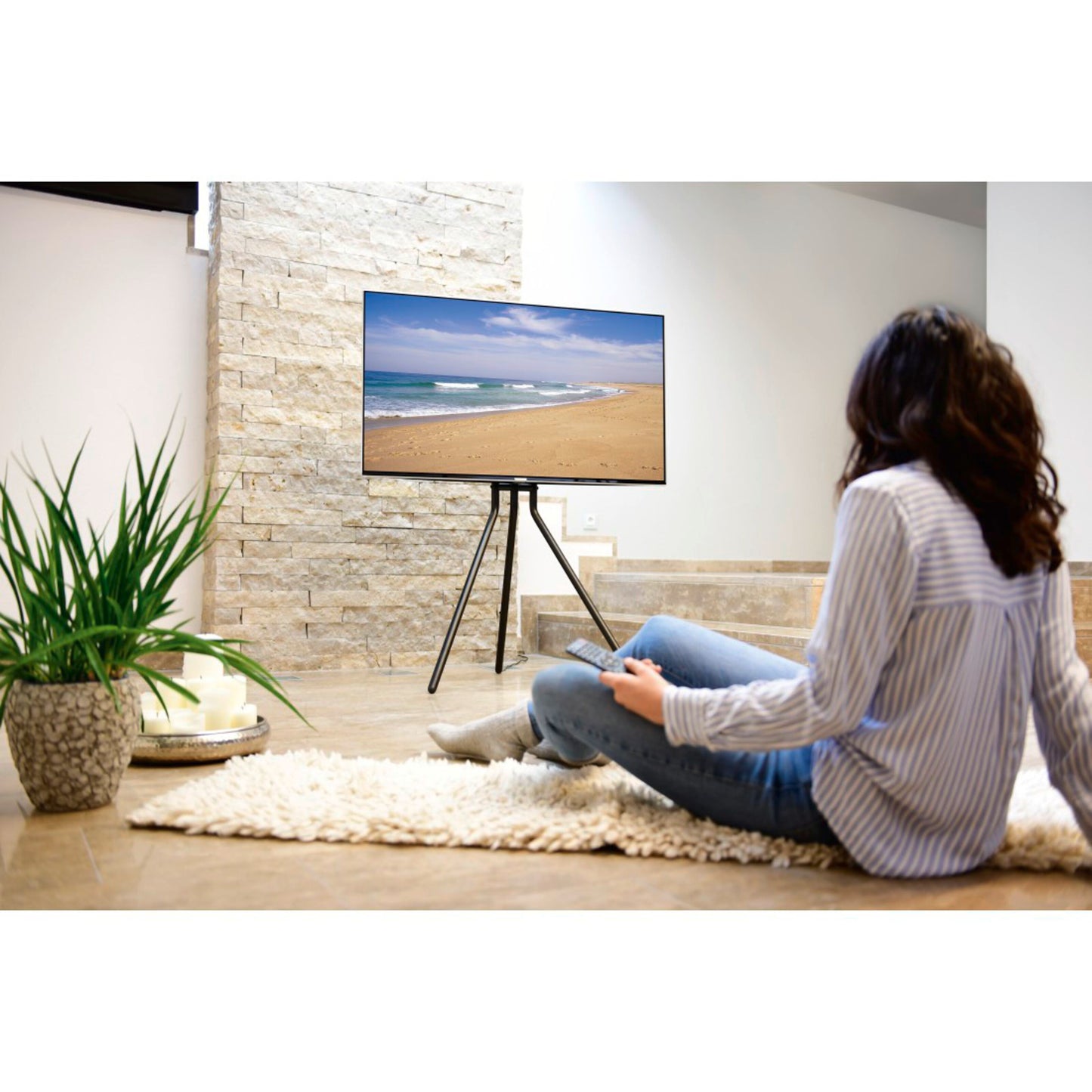 TV på Hama gulvstativ til 37-75" med diskret kabelhåndtering, person foran, tæppe, plante og bakke med kopper.