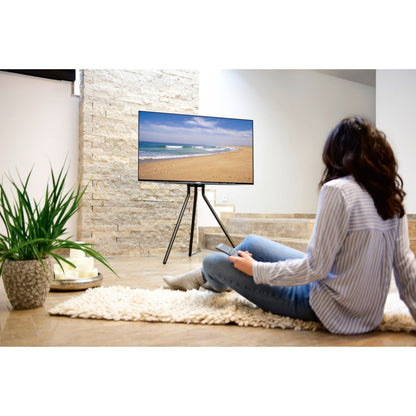 TV på Hama gulvstativ til 37-75" med diskret kabelhåndtering, person foran, tæppe, plante og bakke med kopper.