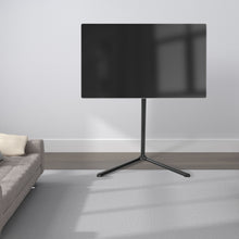 Nikabe V-base gulvstativ sort til tv 49-70 tommer med sort tv, grå sofa og mønstrede puder ses ved siden af.