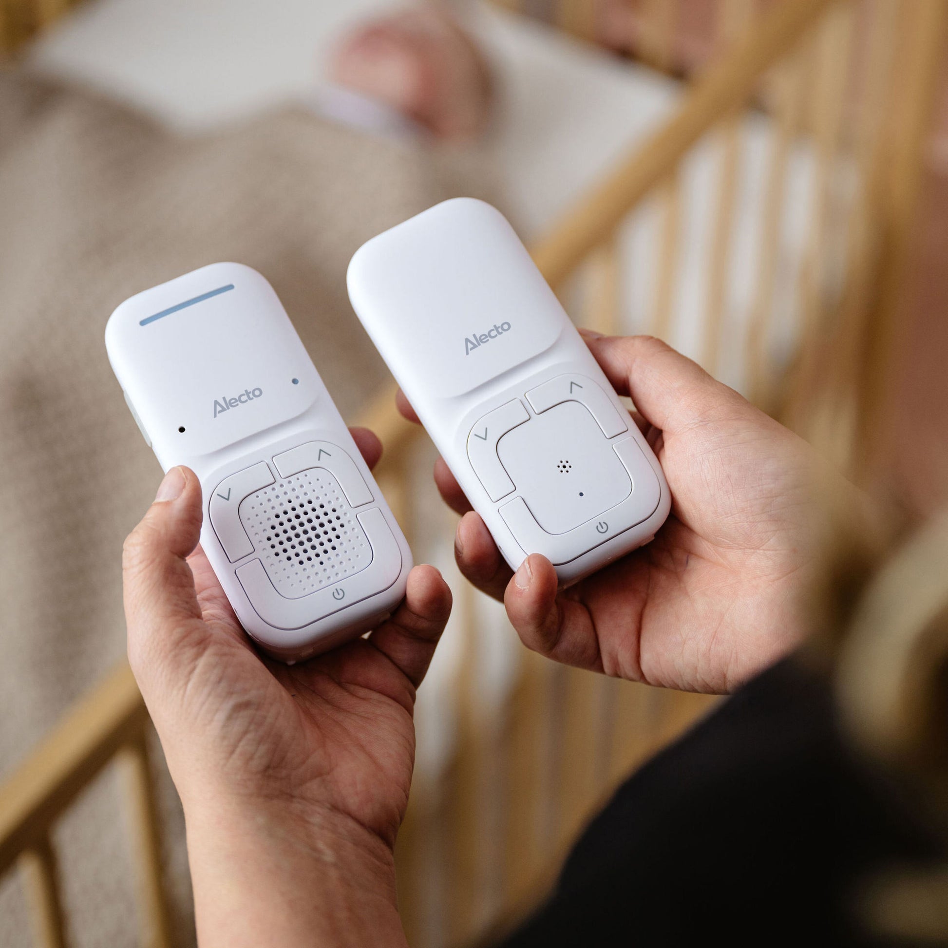 Alecto DBX130 Full Eco DECT-babyalarm, én forældreenhed og én sender i hænder, sløret tremmeseng og babybagved.