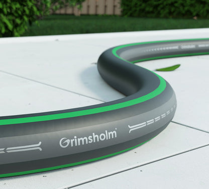 Grimsholm Classic Haveslange 10 m, sort og grå med grøn stribe, fleksibel vandslange nær grønne planter.