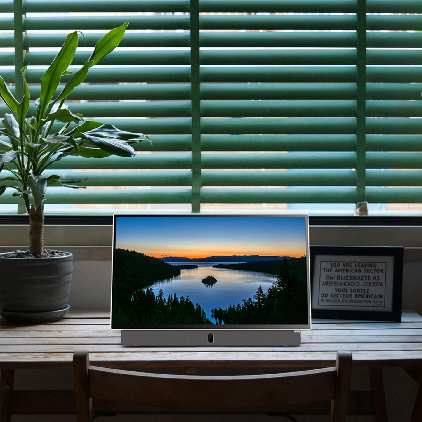 Strong METZ 24" LED bærbar TV på træskrivebord med plante, viser sø og solnedgang, grønne persienner bagved.