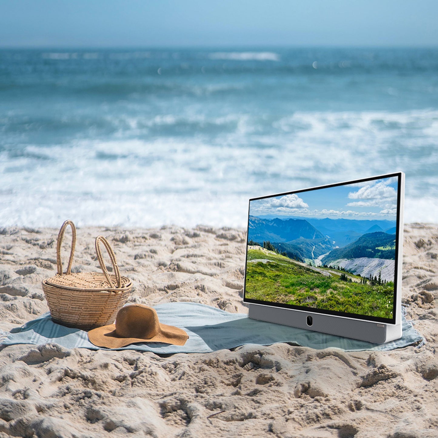 METZ 24" LED bærbar TV viser bjerglandskab, placeret på strand ved siden af stråhat og fletkurv.