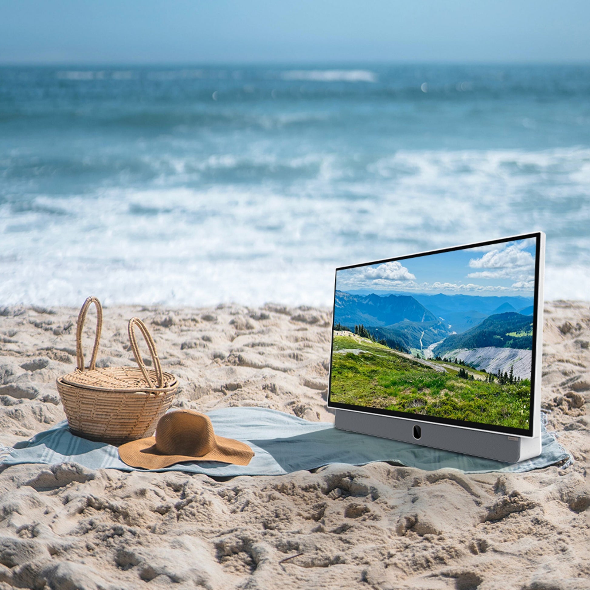 METZ 24" LED bærbar TV viser bjerglandskab, placeret på strand ved siden af stråhat og fletkurv.