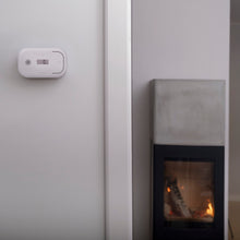 Housegard kuliltealarm med LCD-display på hvidt væg nær pejs med synlig flamme i lyst rum.