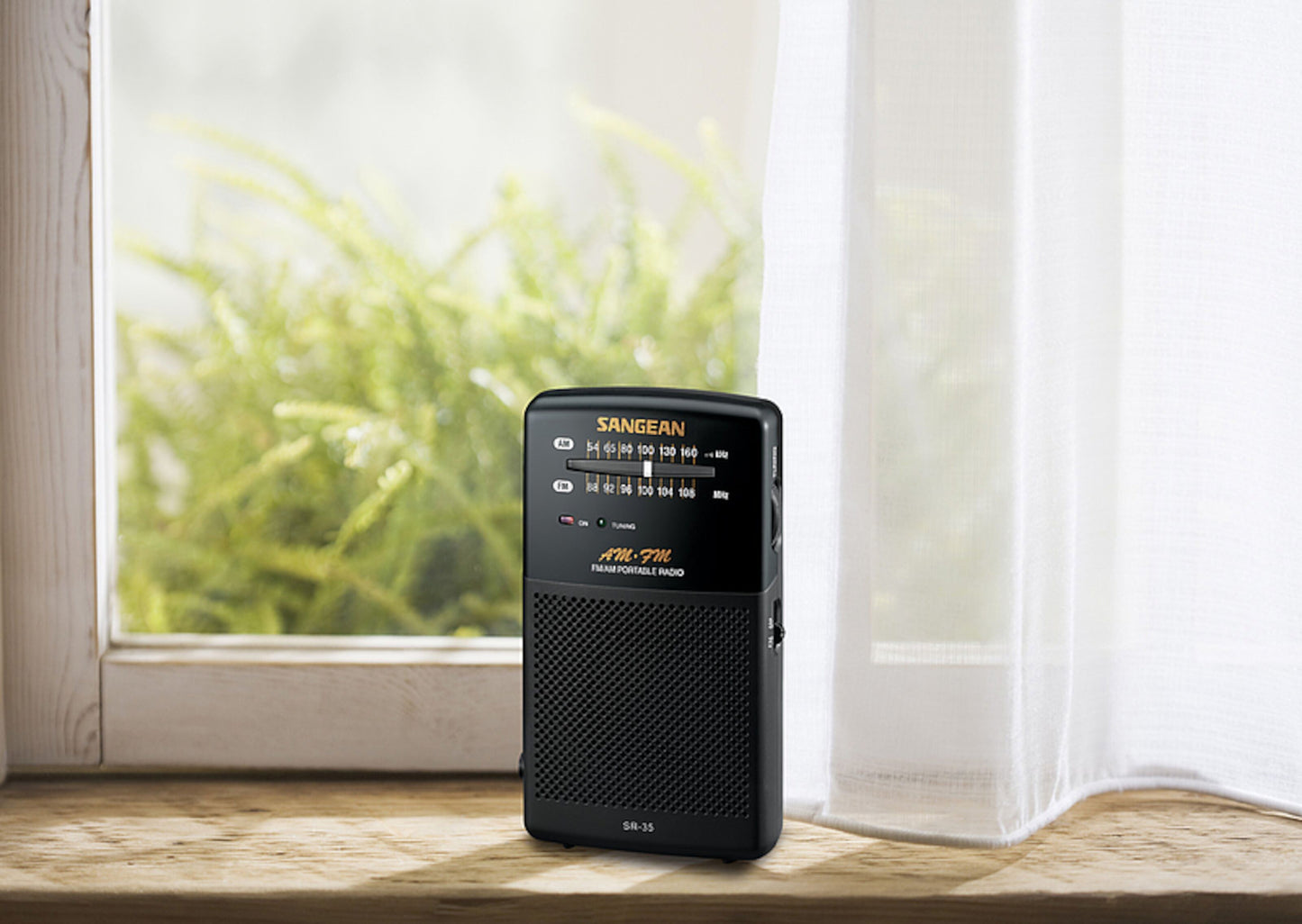 Sangean SR-35 sort FM-radio på trævindueskarm ved hvidt gardin, grønne planter synlige udenfor.