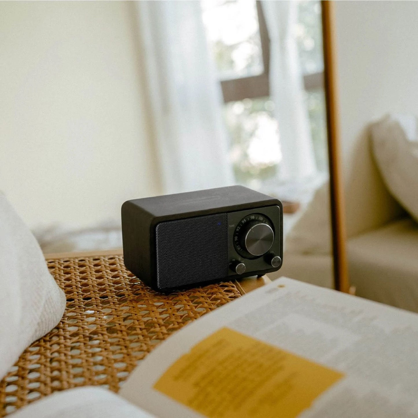 Sangean WR-7 PLUS Midnight Oak FM-radio på fletbord ved bog og hvid pude, synlig front med knapper og display.
