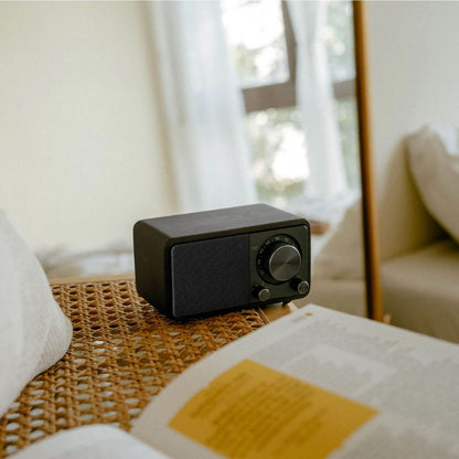 Sangean WR-7 PLUS Midnight Oak FM-radio på fletbord ved bog og hvid pude, synlig front med knapper og display.
