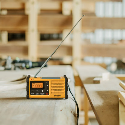 Sangean gul håndsvingradio til FM-signal med USB-C, antenne slået op, sat på træbord i værksted.