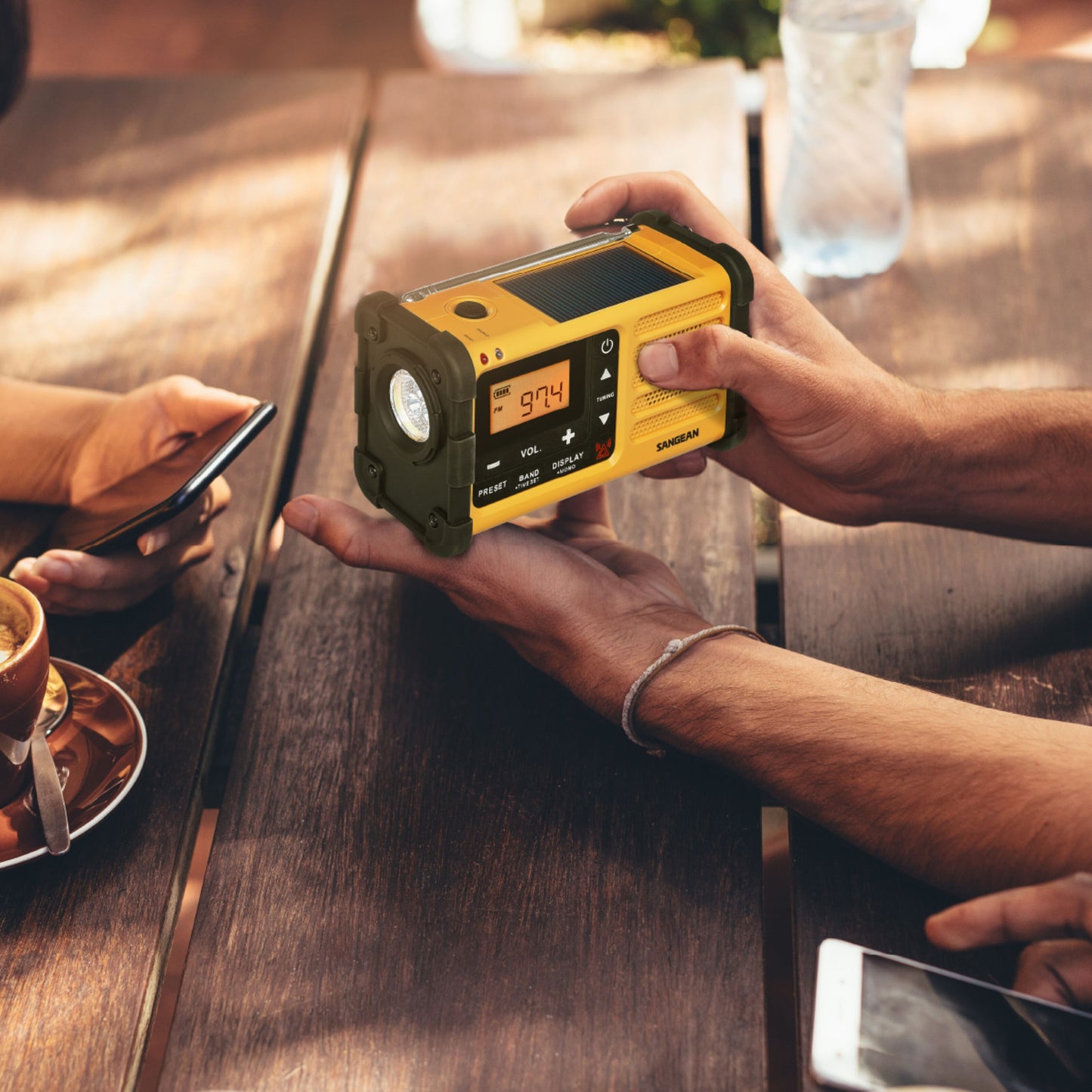 Sangean håndsvingradio FM USB-C holder i hånden, smartphone ved siden af, kop kaffe og vandflaske på bord.