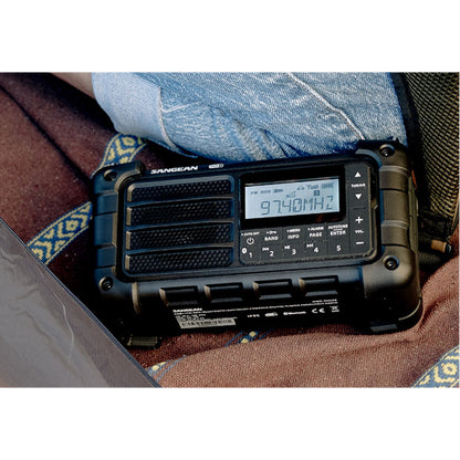 Sangean MMR-99 DAB+ hånddrevet radio med solceller på mønstret stof ved person i denim shorts.