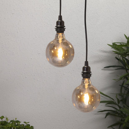 Lampeholder E27 med ledning, synlig filamentpære, ophængt mod hvid væg, grønne planter nederst.