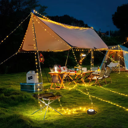 Luxorparts Bærbar campinglygte med LED-lysliste lyser under telt, omgivet af stole, borde og udstyr på græs.