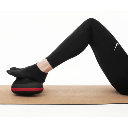 Flowpillow Varmemassagepude på måtte, sort/rød balancepude under fødder, person iført sorte leggings og sokker.
