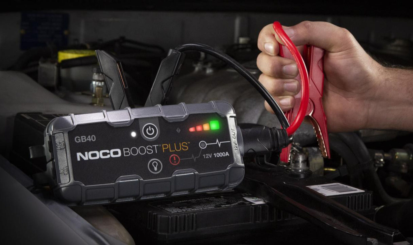Noco Boost Plus GB40 starthjælp, rød kabelklemme til bilbatteri under motorhjelm, hånd holder klemmen.