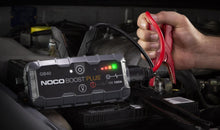 Noco Boost Plus GB40 starthjælp, rød kabelklemme til bilbatteri under motorhjelm, hånd holder klemmen.